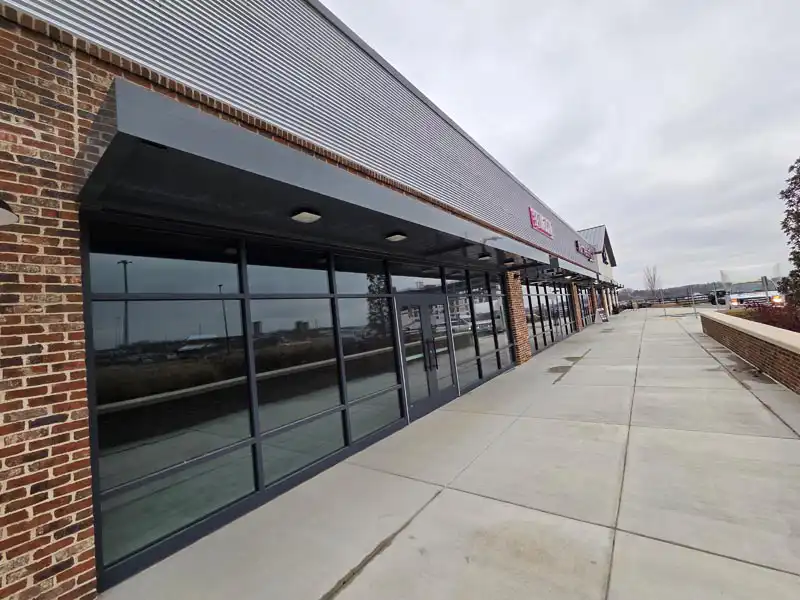 Empty retail storefronts with floor-to-ceiling windows and a wide sidewalk along a commercial strip center.