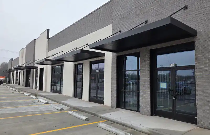Row of modern retail storefronts with glass doors, black awnings, and a parking area in front.