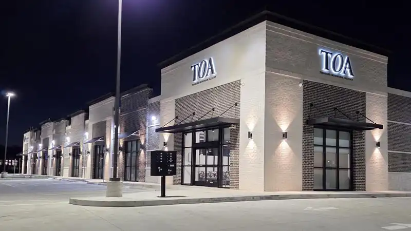 Nighttime view of a retail plaza with illuminated signage, exterior lighting, and multiple storefront entrances.