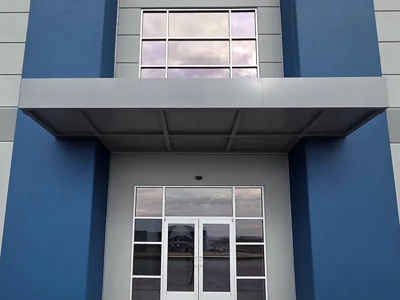 Modern commercial entrance with blue exterior walls, flat metal canopy, and glass double doors beneath a second-story window.