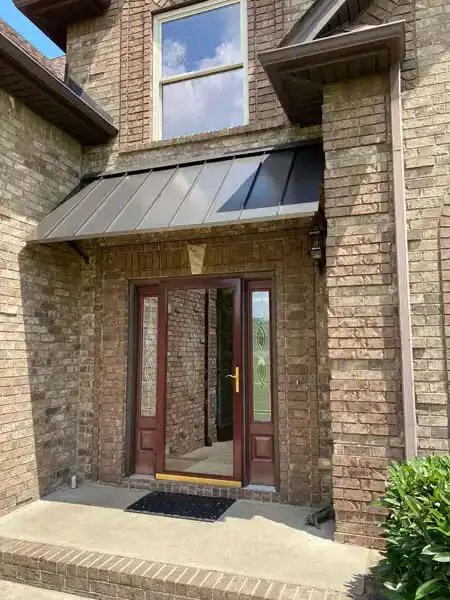 Metal entry awning installed above a glass residential front door surrounded by landscaping.