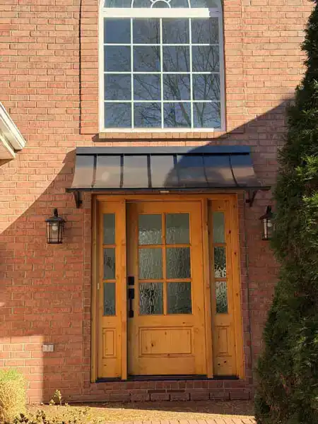 Straight standing seam metal awning mounted above a wooden front door on a brick home.